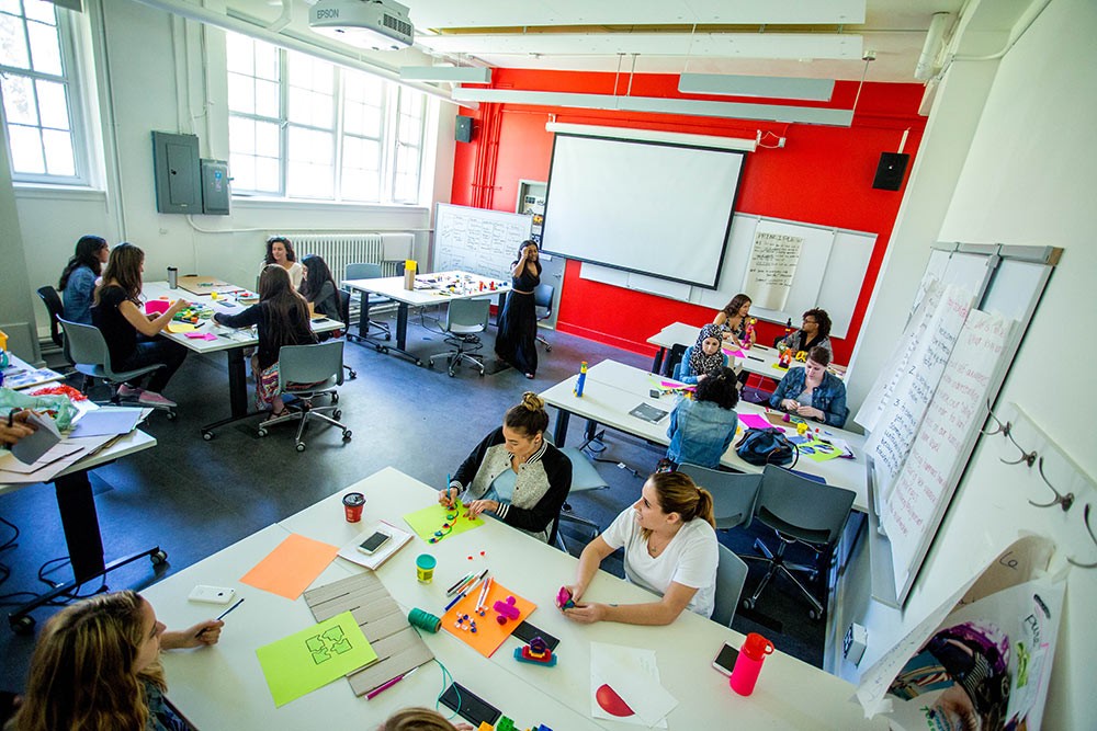 A dozen adult learners sit in small groups around tables with an assortment of creative materials such as coloured paper, markers, post-it notes and modelling clay.