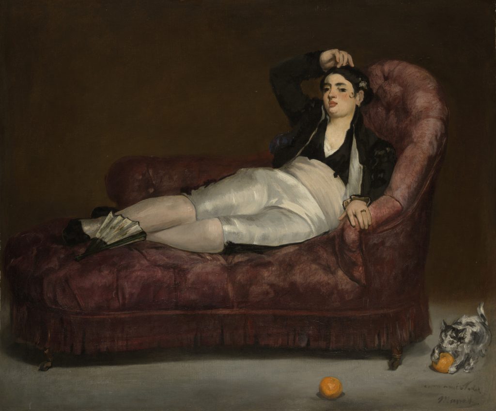 A woman, clad in spanish clothes, reclines on a velvet sofa. On the floor, a small cat claws at an orange.