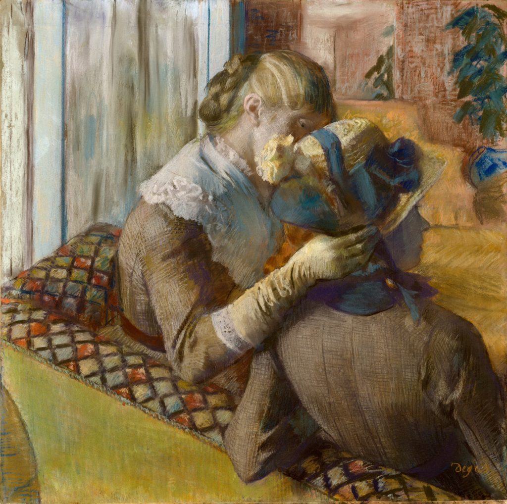 A woman aids another with the trying of a hat, tightened around her chin with a blue bow. They are both seated on a garishly patterned sofa.