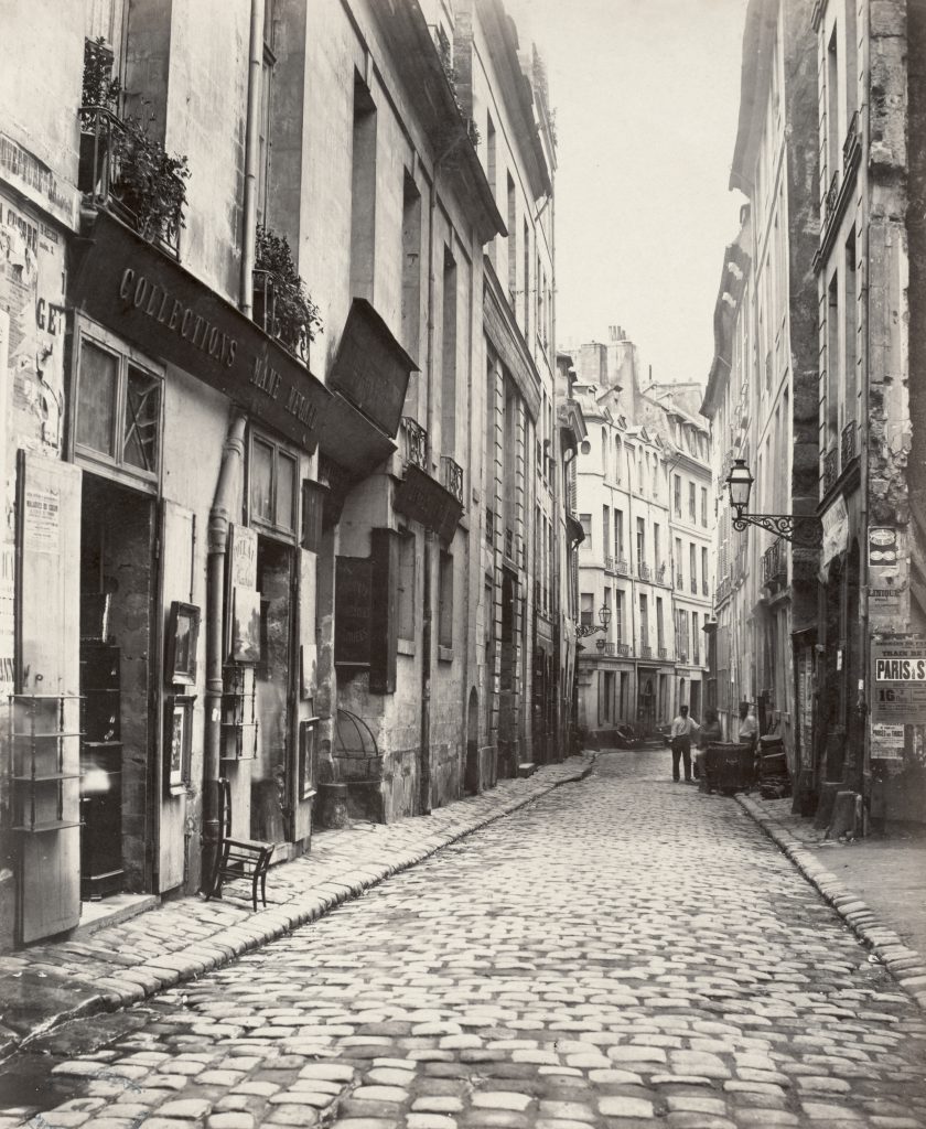 A photograph of a cobblestone street flanked by french shops. A pair of men stand some meters forward.