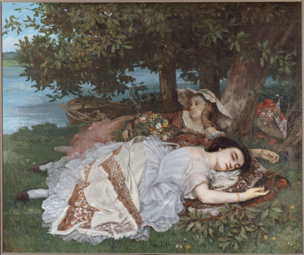 Two young women in dresses lie by the river. They,re surrounded by greenery, a boat lays by the water.