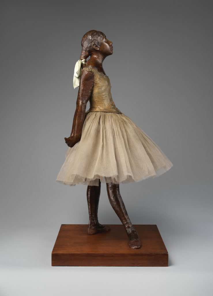 A metallic recreation of a child ballet dancer, in the starting position of a dance. Her costume is coloured yellow faintly.