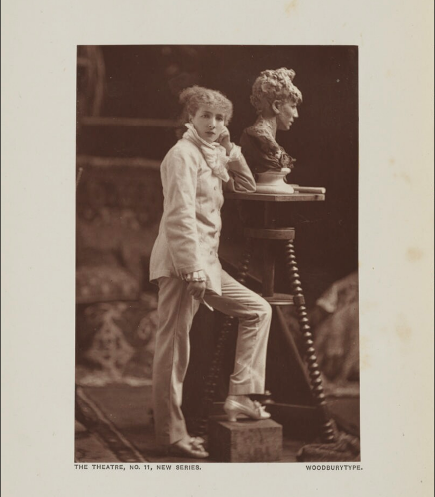 A woodburytype of the young actress on stage, costumed in a formal suit, leaning on a table holding a bust of a woman.