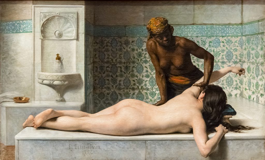 A nude pale-skinned woman lays face-down as a dark-skinned servant massages her arm and back. The servant wears cloths.