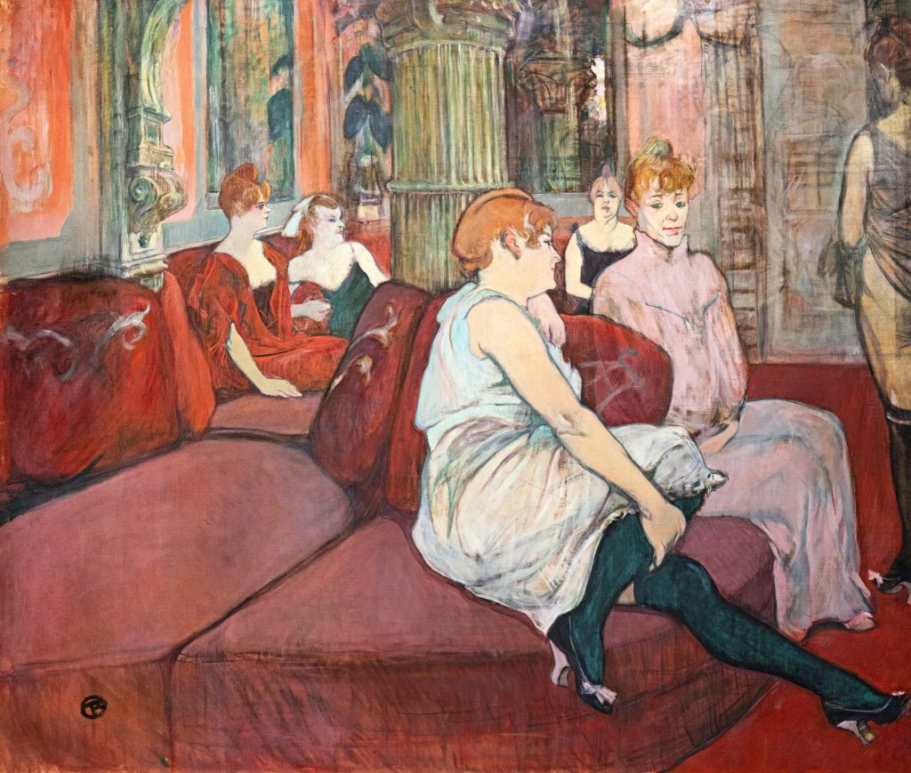 Seated on red sofas across a rather opulent salon are pale women in dresses. Some retain a straight posture while others lean on the furniture.
