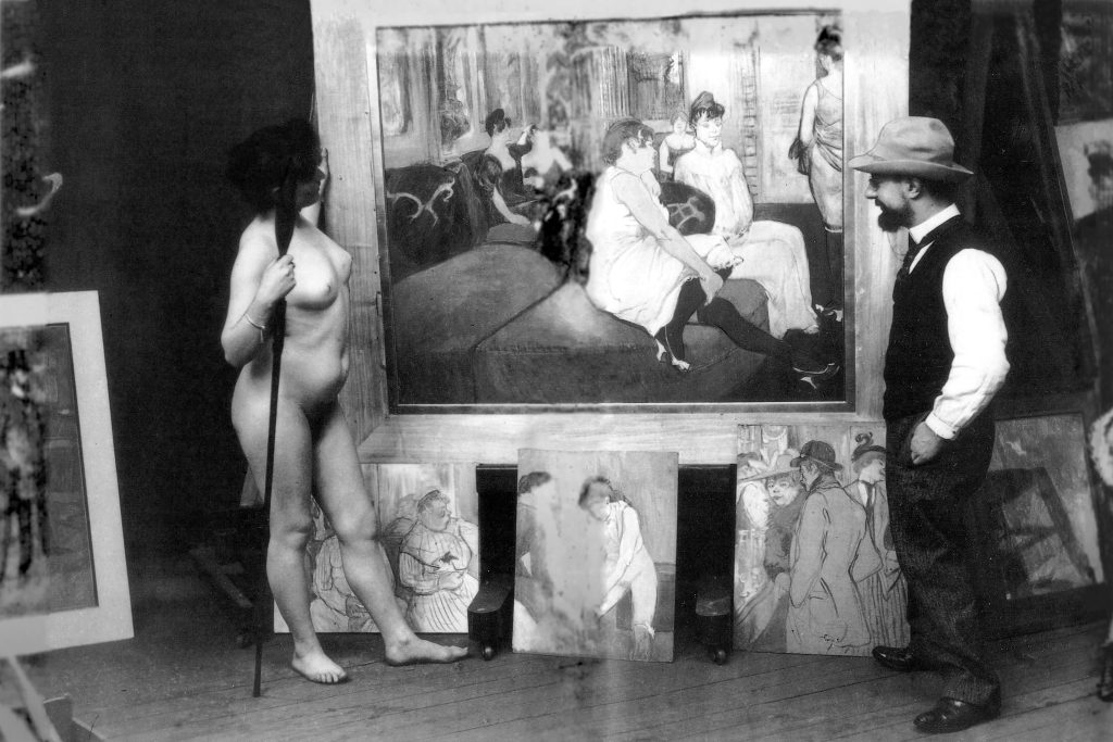 This photograph places Tolouse-Lautrec, dressed in a vest and wearing a pale melon hat, accross a woman standing in the nude. Between them, a framed 'At the Salon of the Rue des Moulins'.