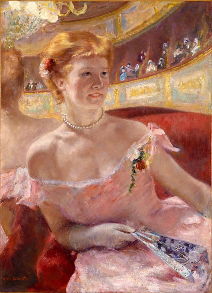 A seated strawberry blonde woman, in an intricate pink dress, delicately holds a fan and leans against a mirror.