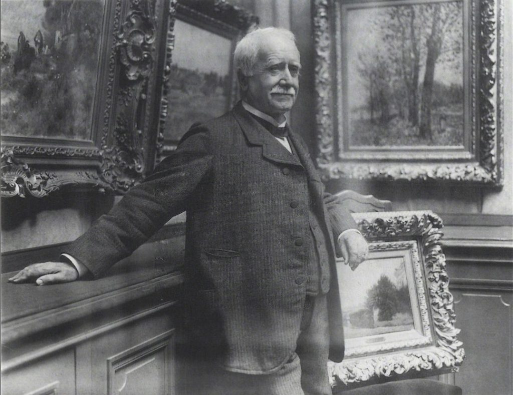 A photograph of Durand-Ruel before a series of paintings in the corner of a gallery. he wears a pinstriped suit and sports a moustache.