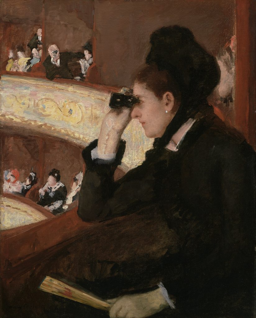 In the lodge balcony of a theatre, a spectating woman leans forward and observes through a set of binoculars.