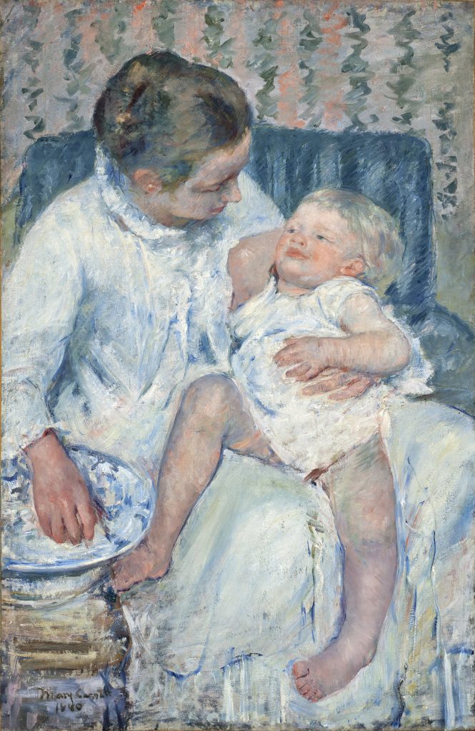 A mother cleans her child on a sofa, her hand drawing from a water basin. Their gazes are inter-locked.