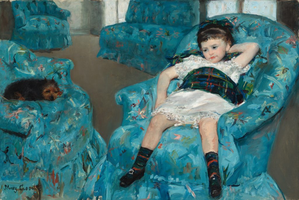 In a room filled with blue patterned furniture, a young girl in a dress lays in an armchair. A dog rests on a sofa by her.