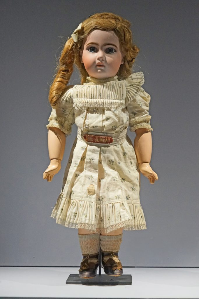 A stiff porcelain doll made to look like a little girl is upright and on display.