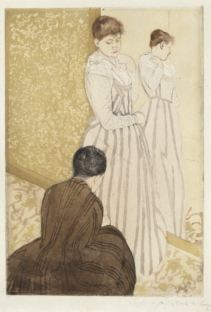 A woman poses in the skeleton of a gown while another woman, clad in black and on her knees, tends to the garment.