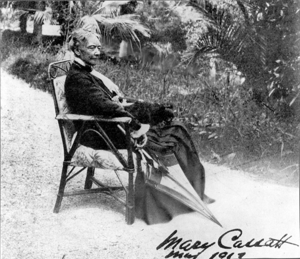 A photograph of a now old Cassatt rests in a chair by a large garden. She holds an umbrella.
