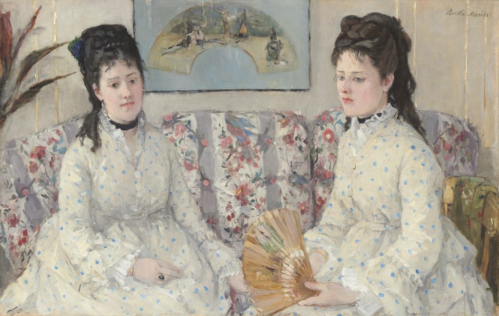 Two women in lace white dresses sit on an ornate couch, one sorrow in expression. On the wall behind them is an art-work.