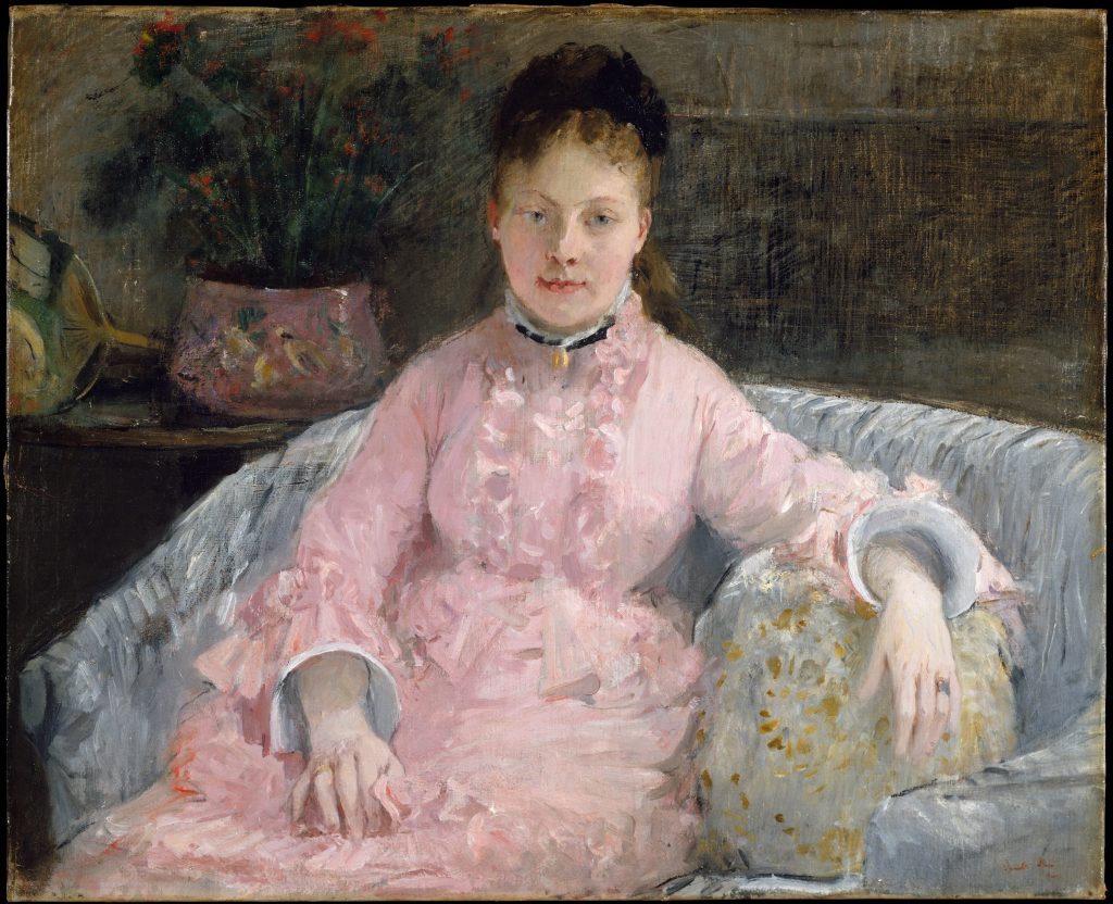 In a pink dress and a black head-piece, a woman rests on a couch with one arm posed over a cushion.