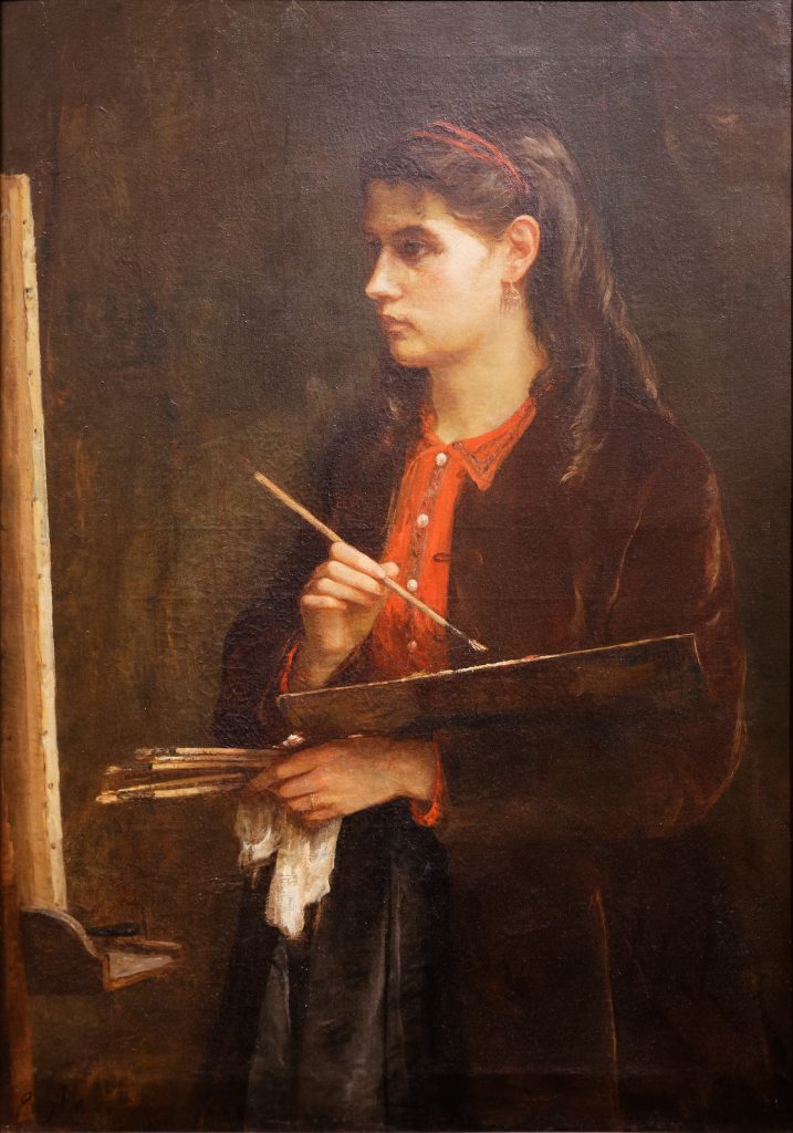 A portrait of Morisot painting, standing by a canvas. A brown over-coat covers a red dress-shirt, and her hair is done into a long pony-tail.
