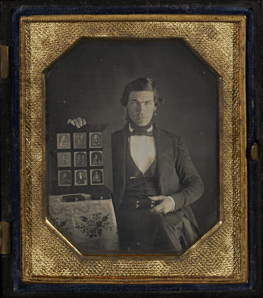 A photo of a suited man holding a display book of daguerreotype photographs and a wooden box, presumably a daguerreotype.