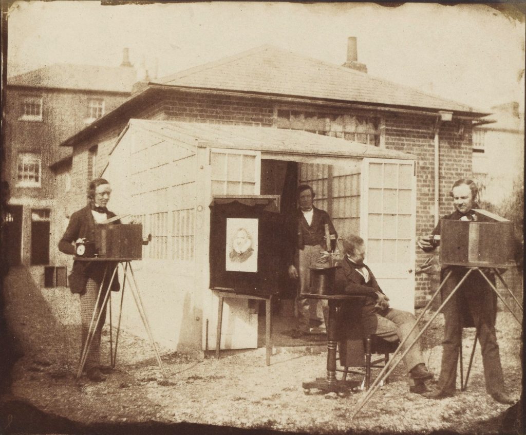 A small group of men setting up photographic devices by a shed.
