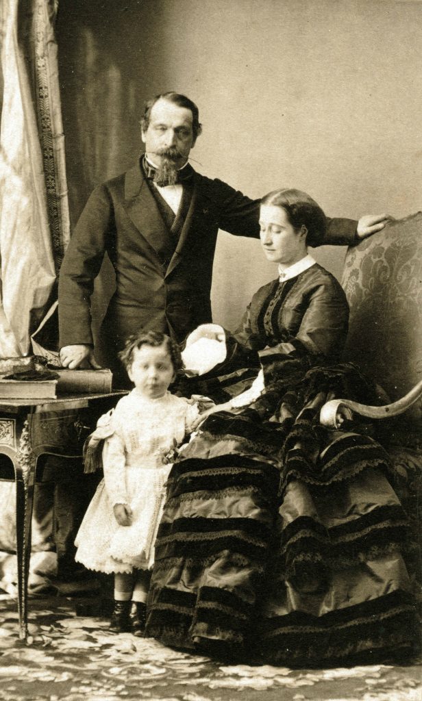 A silver printed family portrait in a makeshift living-room setting. The patriarch of the family, Napoléon III, stands above his wife and child.
