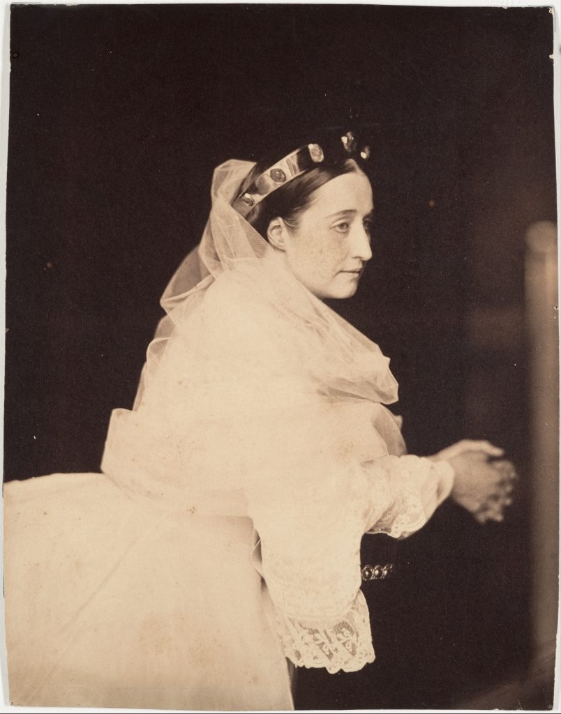 A portrait of a woman in a white dress, set before a dark backdrop, bearing a crown on her haid.