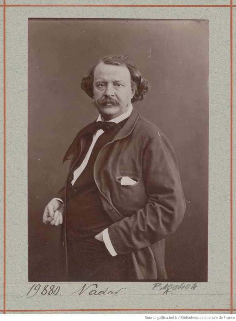 A silver print portrait of a suited man, eyes forward, leaning slightly on his cane. His clothing is dapper and he sports a large moustache.