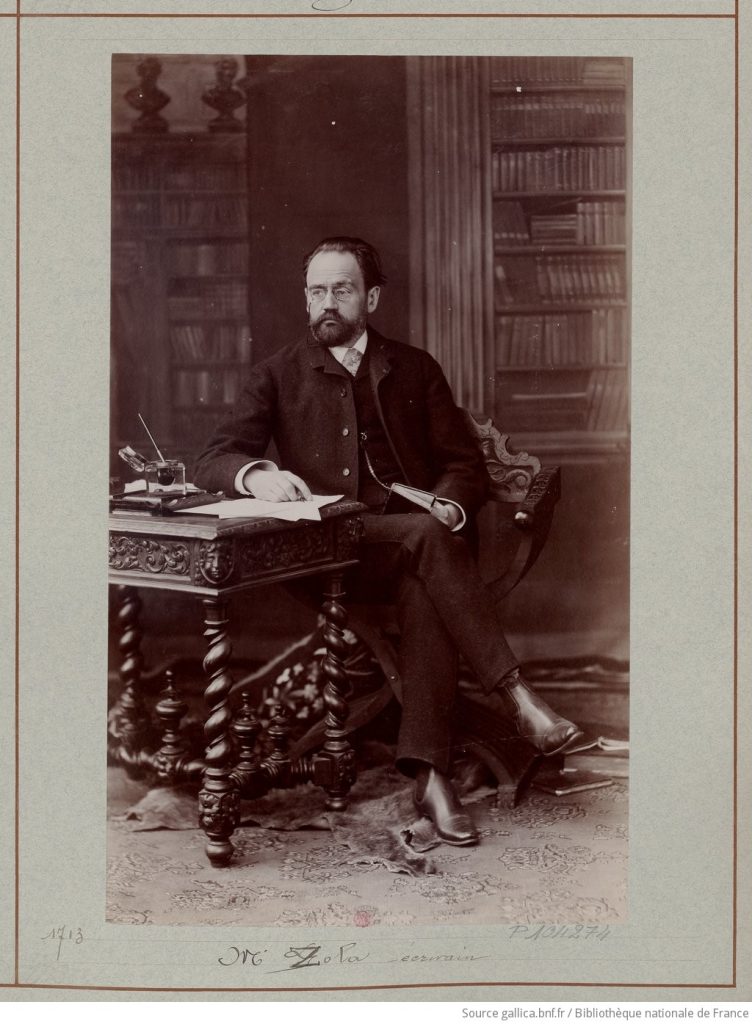 Silver print photograph of the writer Émile Zola, sat at an ornate wood desk, before a backdrop of library aisles.