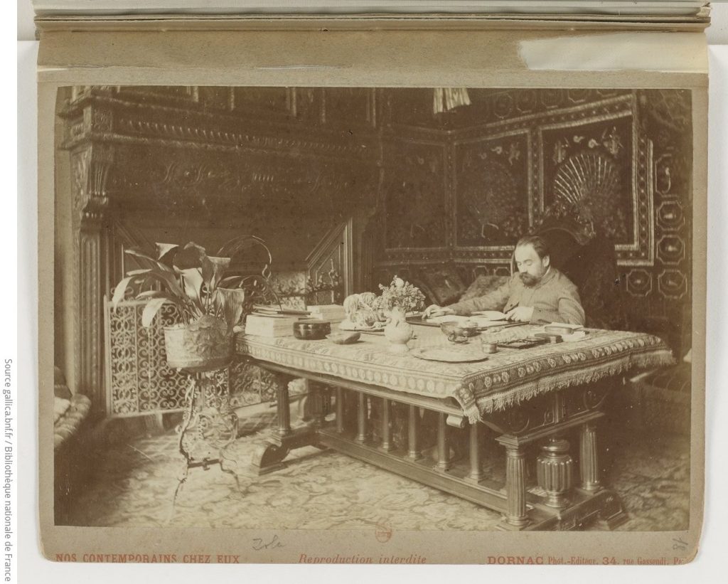 A posed beige tinted photograph of Zola sat a vast desk littered with manuscripts and dining sets.