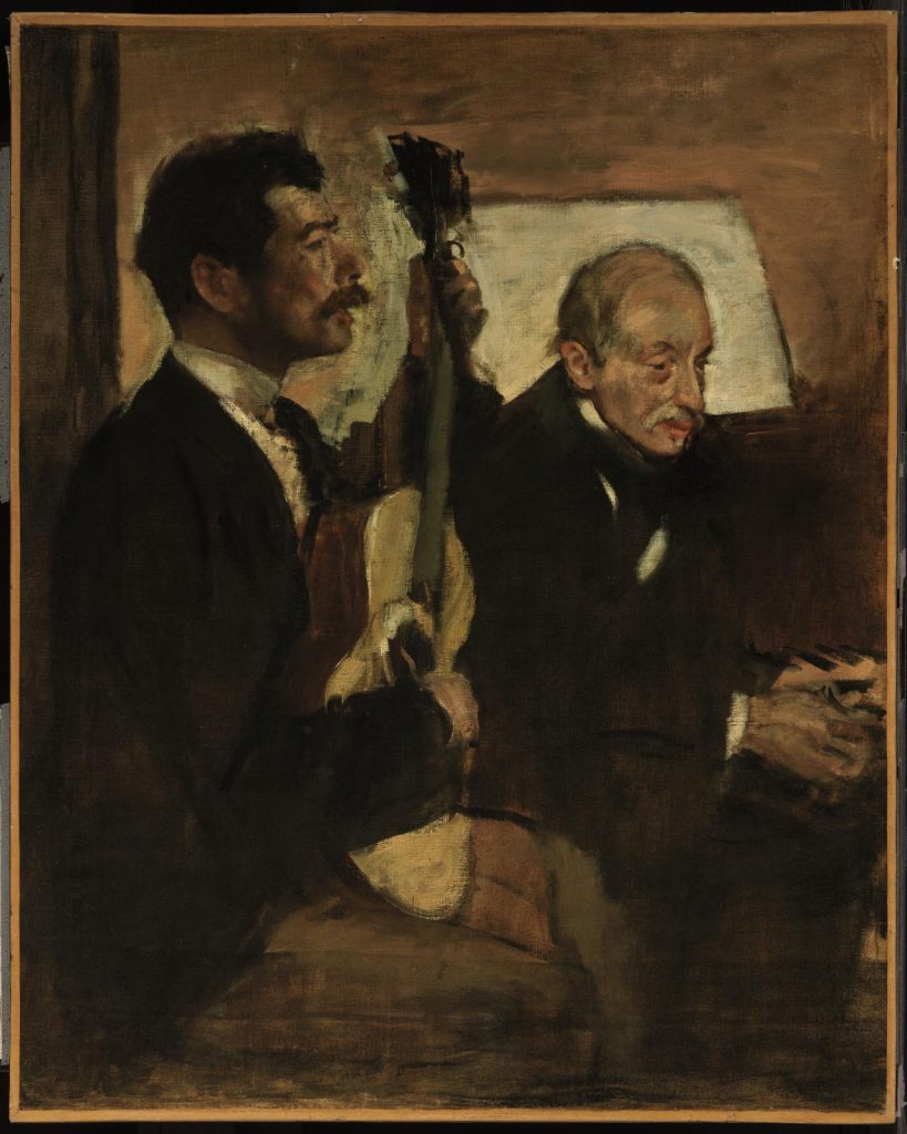 An older man leans forward, attentively listening to the guitarist sitting next to him. A dark but warmly lit scene.