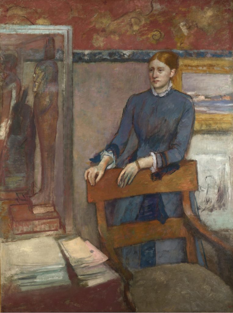 A blue dressed woman stands behind the chair of an office study, where paintings and canvases are hung.
