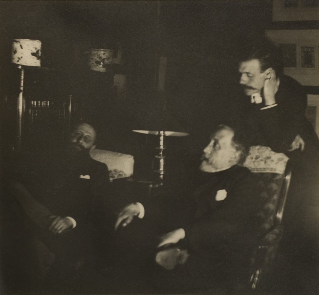 A very dark photo of three men, two seated and one leaning on the former's armchair, in somber contemplation within a room.