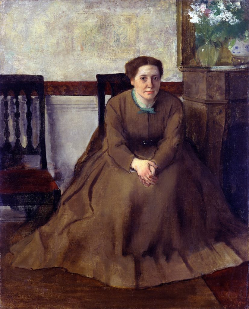 A seated woman in a brown dress leans forward, hands clasped. By her is an art-work and flower arrangement.