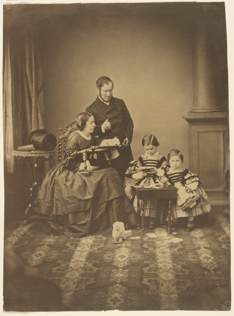 A family silver print portrait, the father stands above his sitting wide and their two unfiformly dressed daughters. They all pose pre-occupied.