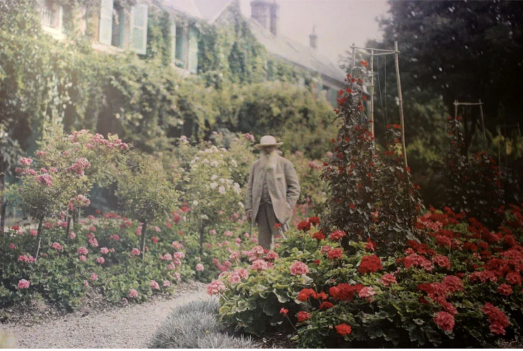 A photograph of a bearded Monet standing in his luscious estate garden.