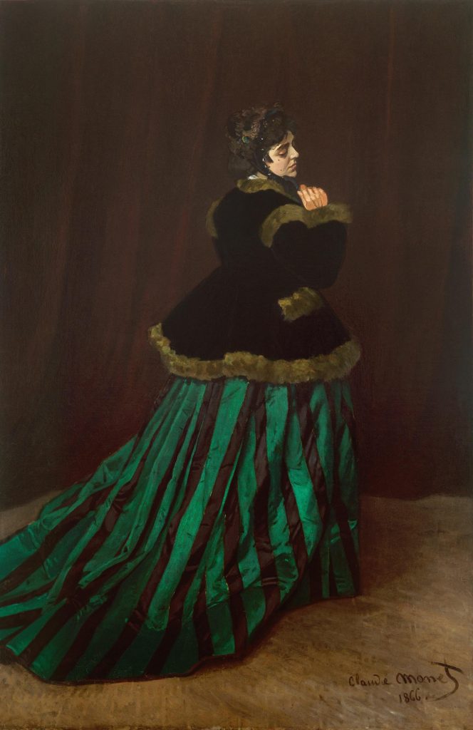 Before a brown curtained backdrop, we observe a woman in a flowing green dress from behind. The light catches her robes and her face, shyly turned towards us, is clutched slightly by her right hand.
