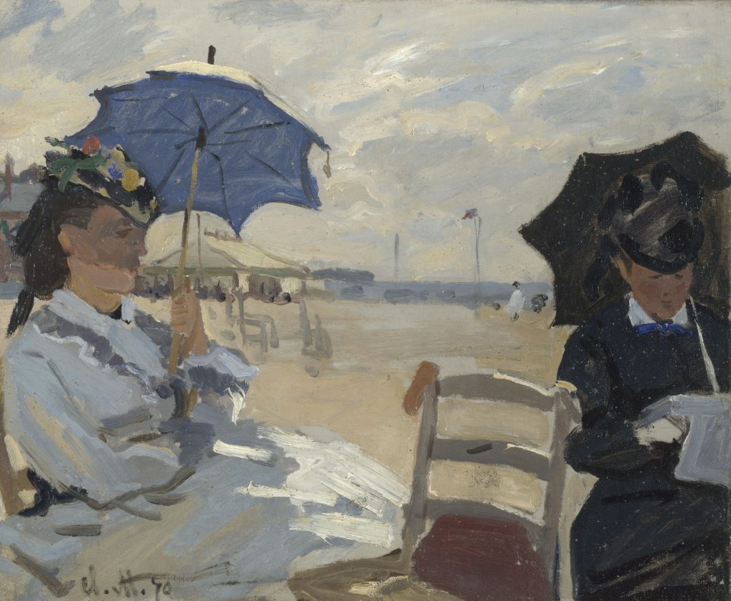 Two woman, one in a beige dress and one in black, sit on the beach. One reads the journals, the other observes.
