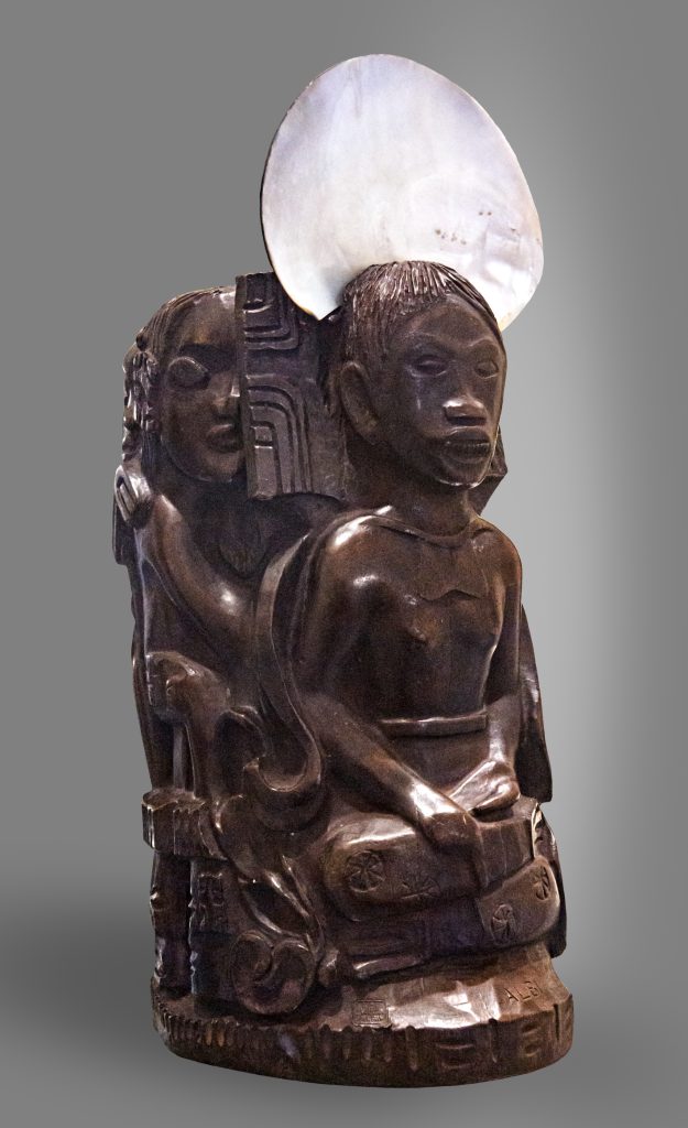 A carved wood sculpture, mimicking indigenous sculpture figures. A shell is embedded into the head of the piece.