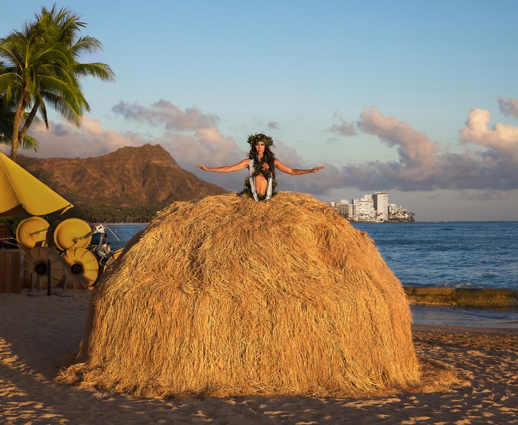 A posed stereotypical hula girl, her straw dress enormously exaggerated. A contemporary photograph.