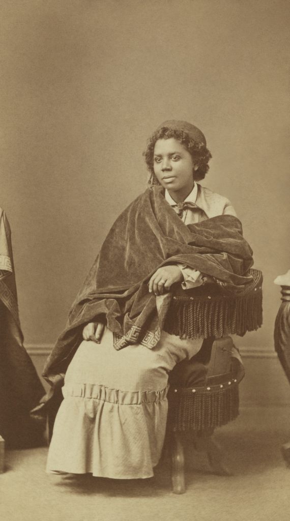 A silver photographic portrait of Edmonia Lewis sat in a chair, a blanket draped over her.