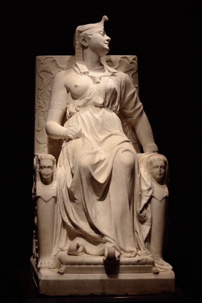 A marble enthroned Cleopatra, bossom semi-exposed, looks to the right as her head rests on the throne's back.