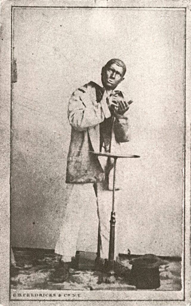 An aged photograph of a white man in blackface wearing a rugged coat and hunching over rythm bones in his palm.