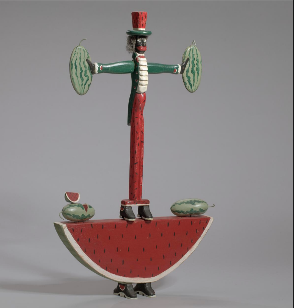 A scultpure of a black caricature anthropomorphized into a watermelon, standing on top of a watermelon slice.