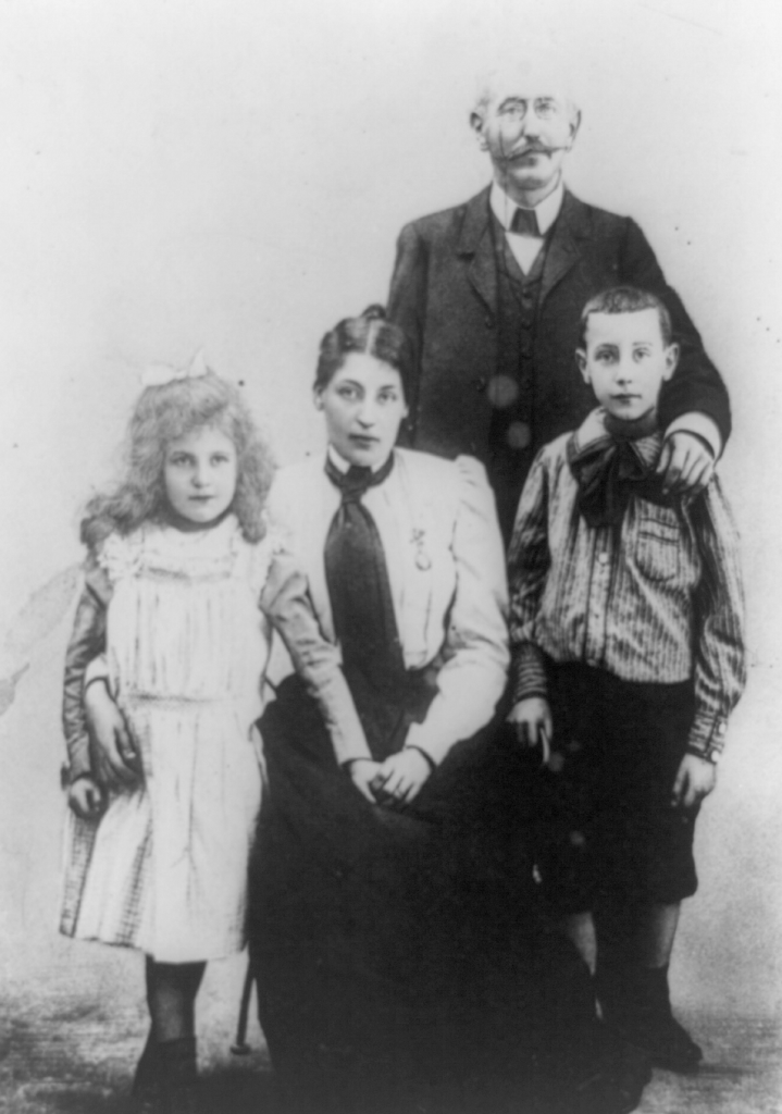 A straight photographic portrait of Dreyfus standing above his wife and children, arms outstretched on their shoulders.