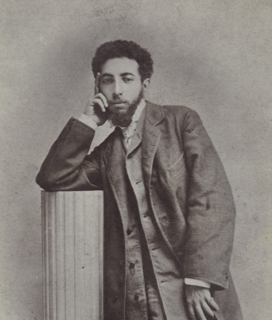 A portrait photograph of Gottlieb leaning on a column prop, fingers resting on his temple. He wears a formal coat and sports beard, he looks forward.
