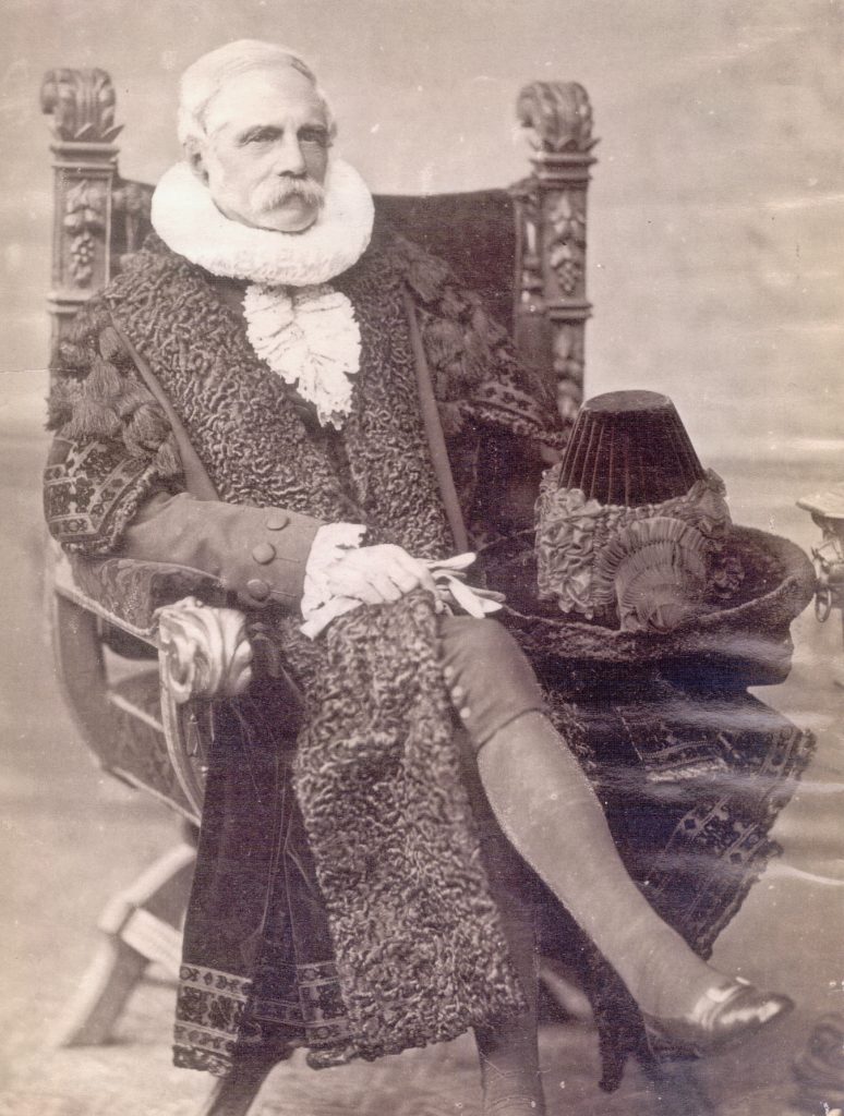 A stoic portrait photograph of Peterson, in an ornate black robe and a very intricate black hat sitting on his lap. He sits in a throne-like chair.