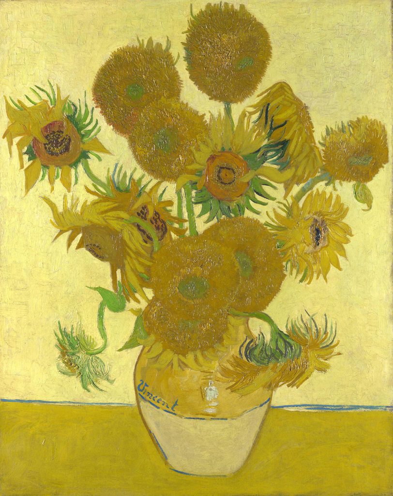 A vase of warm sunflowers placed on a table before a yellow wall.
