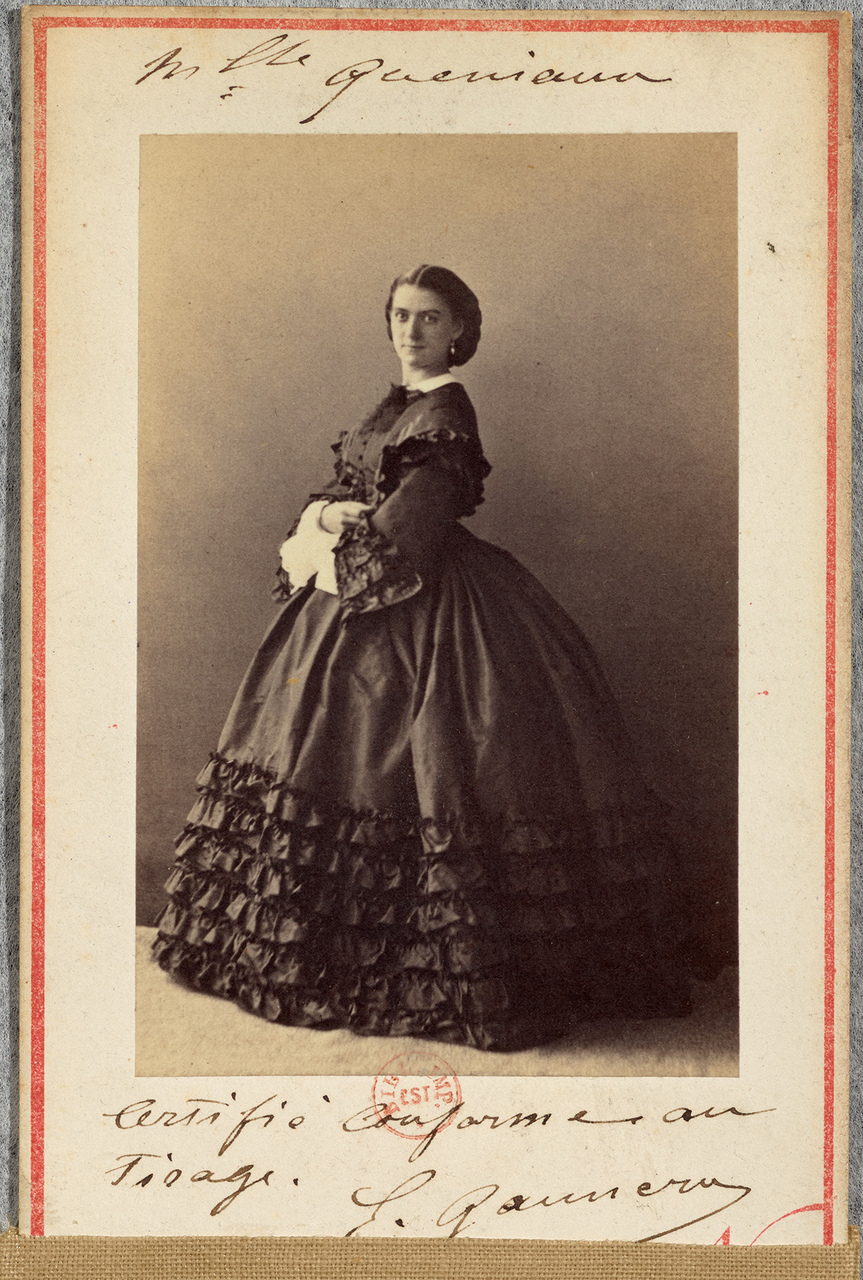 A photograph of a woman, standing, in a formal black dress. Written by the photo's frame is the name of the sitter, 'Mme Queniaux'.