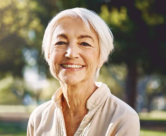A retirement-age white woman smiles against a backdrop of trees.