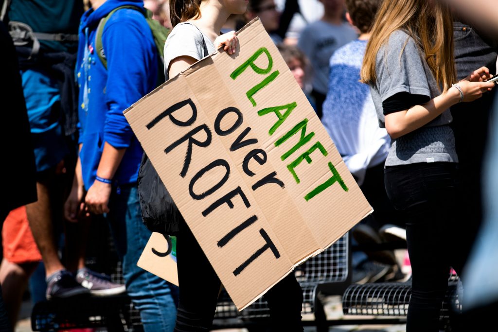 A protest crowd with young adults. A woman is holding a cardboard sign that reads 'PLANET OVER PROFIT'