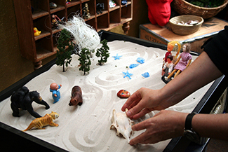 This photograph shows a person playing with objects in a small box filled with sand. The person is organising these objects and small play figures in a form of treatment called sandplay.